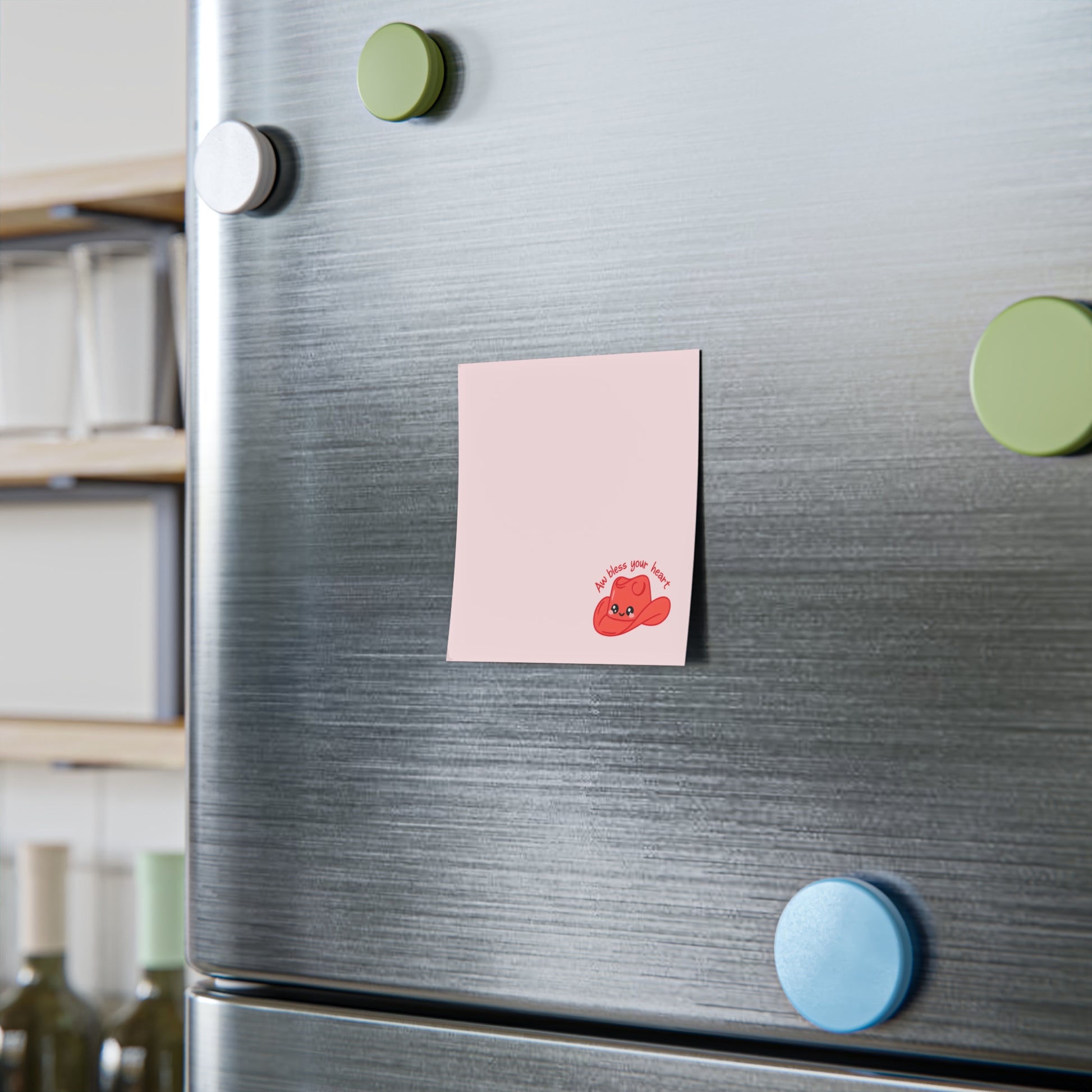 Pink sticky note with a small red cowboy hat character and the phrase “Aw bless your heart” printed at the bottom, attached to a stainless steel refrigerator door with colorful round magnets nearby.