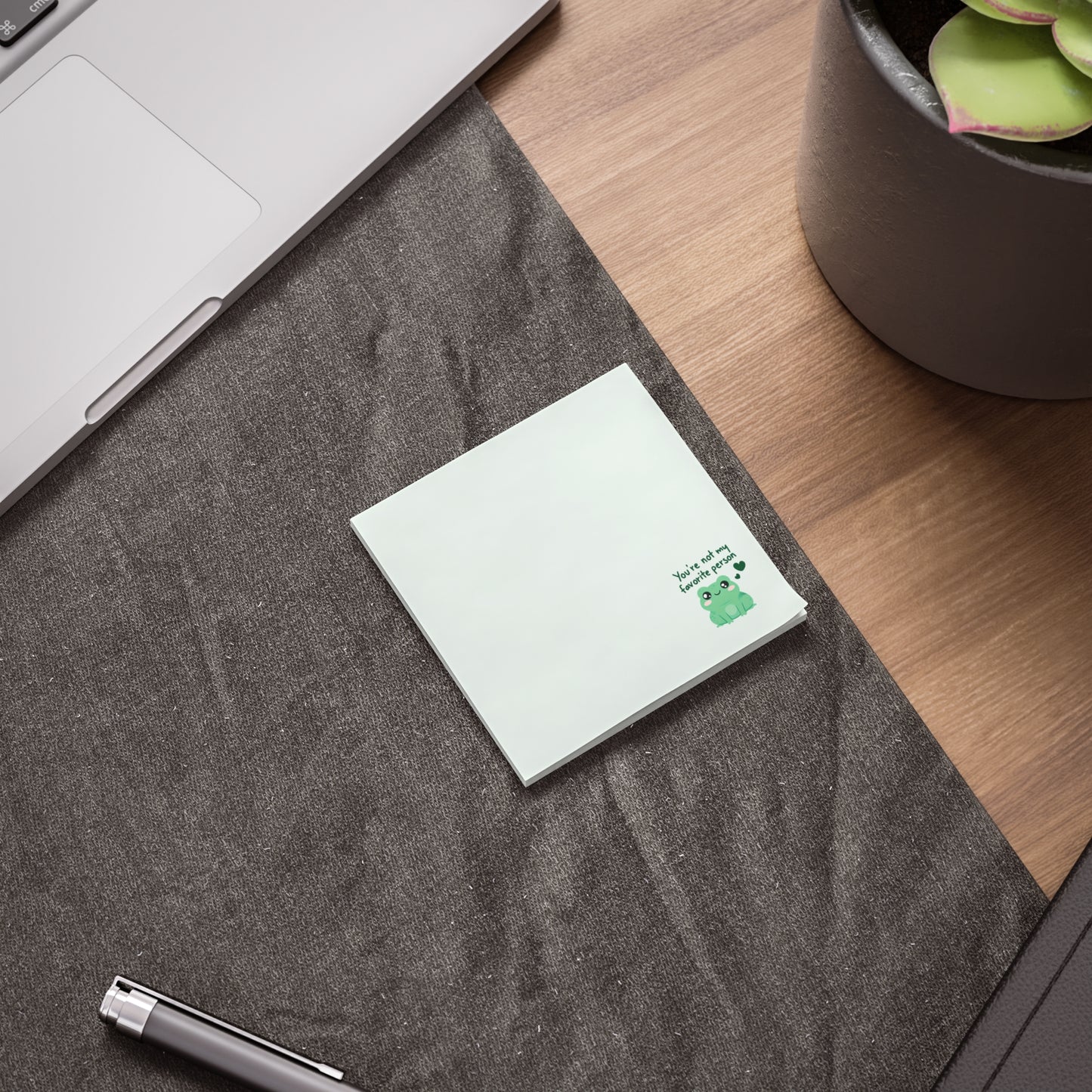 Small white notepad with a green design on a gray textured surface next to a laptop and plant.