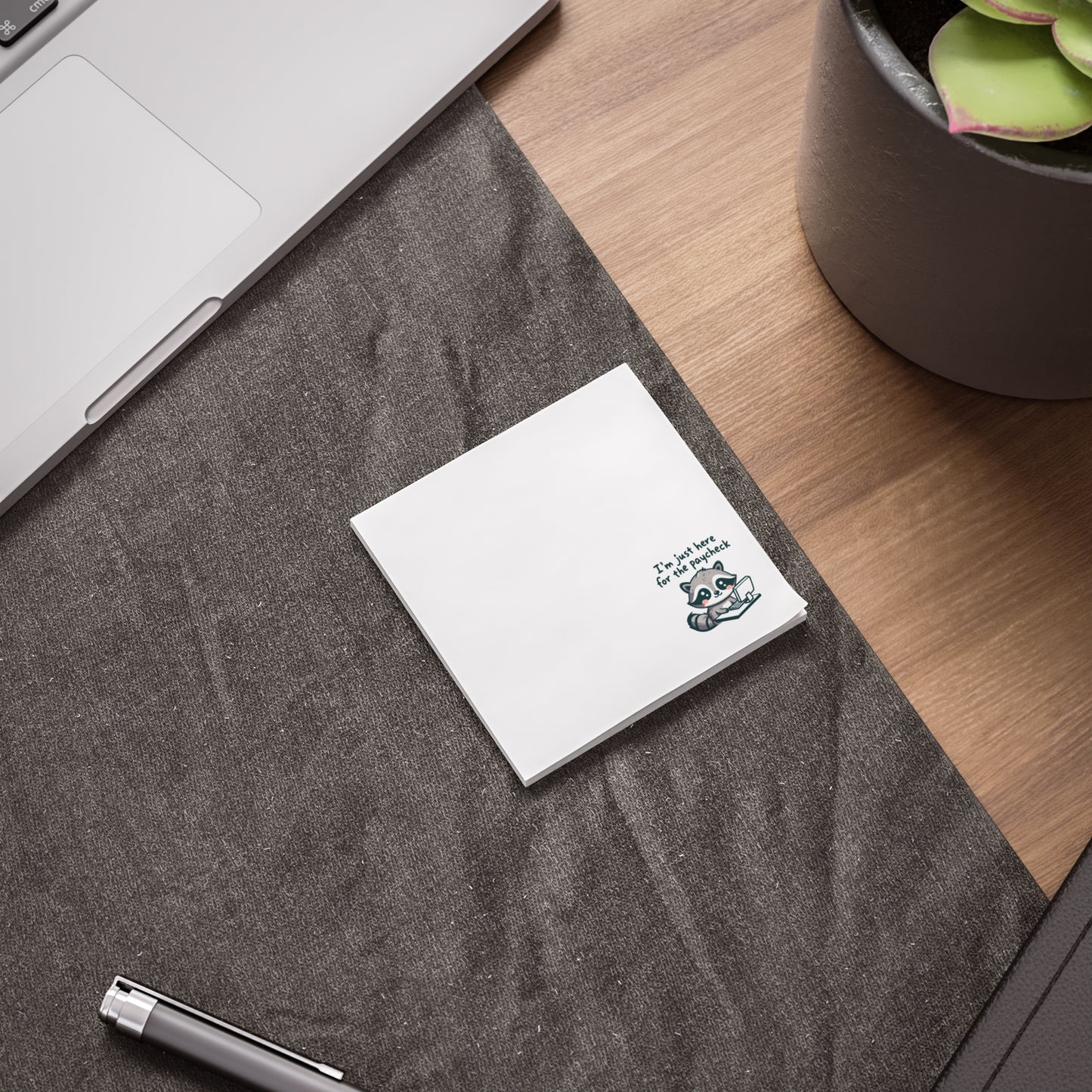 White sticky note pad with the raccoon and “I’m just here for the paycheck” text in the bottom right corner, placed on a dark fabric desk mat beside a laptop, pen, and plant on a wooden desk.
