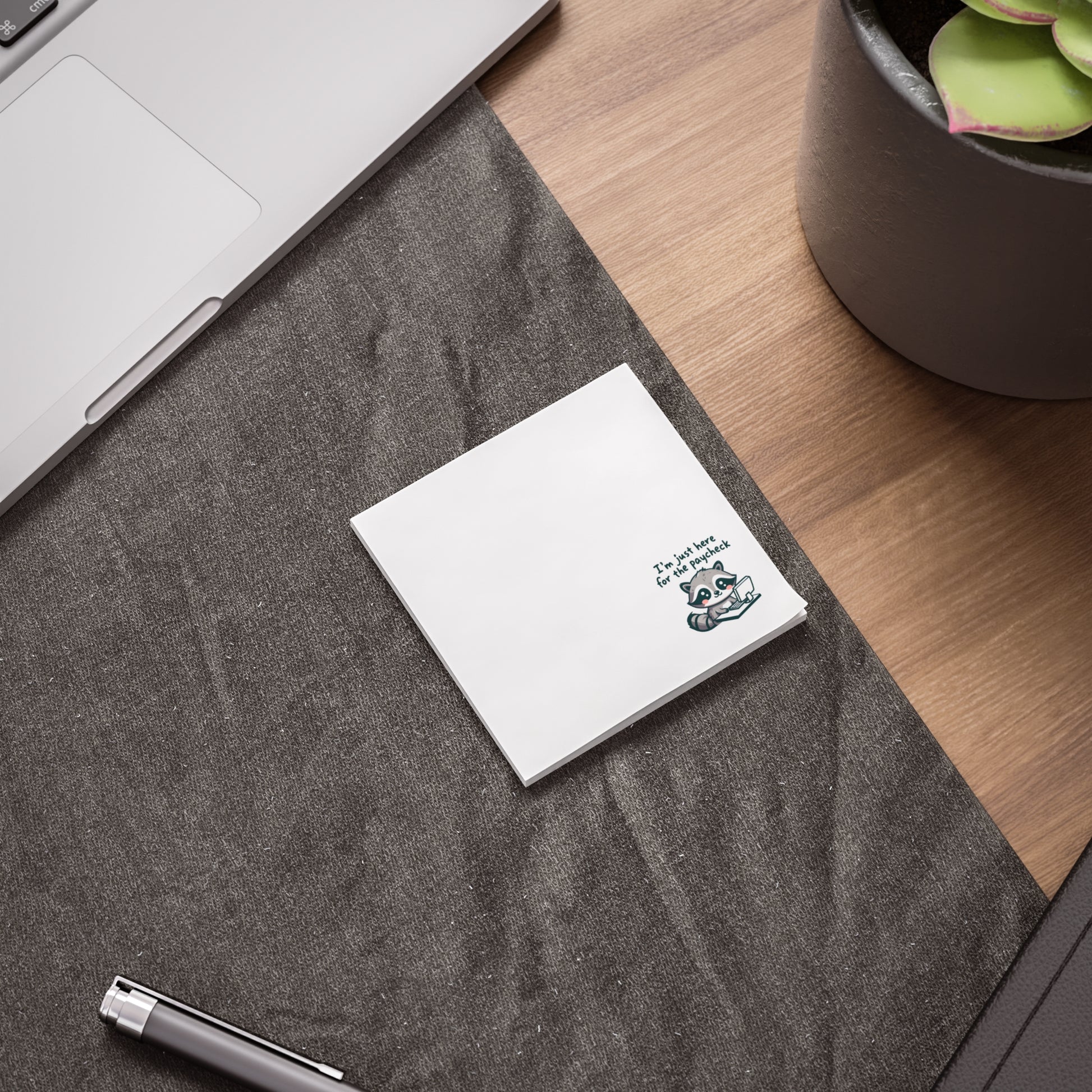 White sticky note pad with the raccoon and “I’m just here for the paycheck” text in the bottom right corner, placed on a dark fabric desk mat beside a laptop, pen, and plant on a wooden desk.