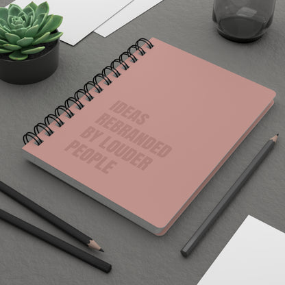 Pink notebook with text on a gray surface with pencils and a plant.